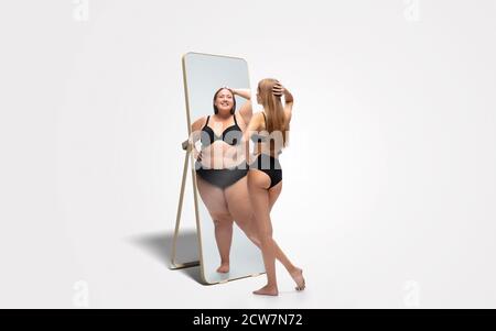 Junge fit, schlanke Frau mit Blick auf fette Mädchen in Spiegelreflexion auf weißem Hintergrund. Sie denkt, dass sie nicht genug sportlich ist. Konzept der gesunden Lebensweise, Fitness, Sport, Ernährung und Körper positiv. Stockfoto