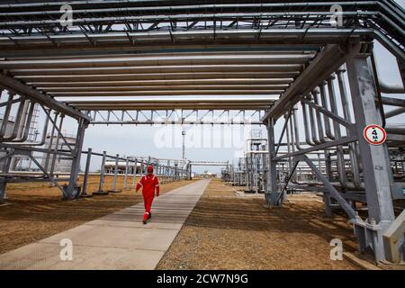 Ölarbeiter in rot Arbeit tragen und roten Helm unter den Rohrleitungen. Ölraffinerie-Anlage in Wüste mit gelbem Sand. Zhaik-Munai Öllagerstätte, Kasachstan. Stockfoto