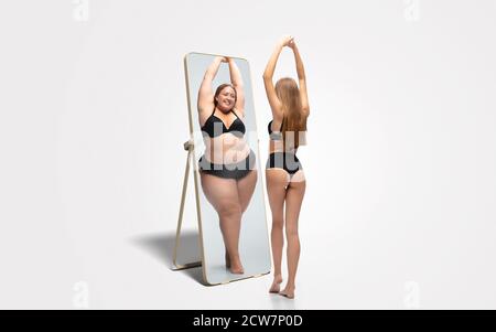 Junge fit, schlanke Frau mit Blick auf fette Mädchen in Spiegelreflexion auf weißem Hintergrund. Sie denkt, dass sie nicht genug sportlich ist. Konzept der gesunden Lebensweise, Fitness, Sport, Ernährung und Körper positiv. Stockfoto