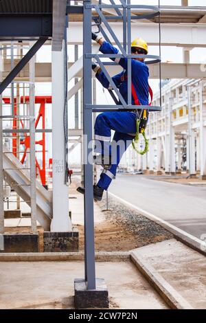 Zhaik-Munai Öllagerstätte, Kasachstan. Ölraffinerie und Gasverarbeitungsanlage. Industriekletterer in blau Arbeitskleidung und gelben Hardhut auf Metalltreppe. Stockfoto