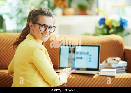 Glückliche elegante Frau in gelber Jacke im modernen Haus in sonnigen Tag gehen, um Webinar auf einem Laptop zu sehen. Stockfoto