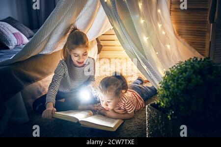 Zwei niedliche kleine Kinder lesen ein Buch mit Taschenlampen im Zelt. Glückliche Mädchen spielen zu Hause. Lustige, nette Kinder, die Spaß im Zimmer haben. Stockfoto