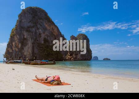 Eine ältere Frau liegt an einem tropischen Sandstrand und sonnen sich. Vor dem Hintergrund von Felsen und Booten im Meer. Stockfoto