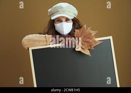 Hallo Herbst. Lächelnd elegante Hausfrau mittleren Alters im Schal mit weißer medizinischer Maske und gelben Herbst Ahornblätter zeigt leere schwarze Tafel auf bronz Stockfoto