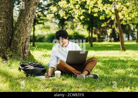 Männlicher Student, der sich Notizen gemacht hat, während er mit einem Laptop im Park sitzt Stockfoto