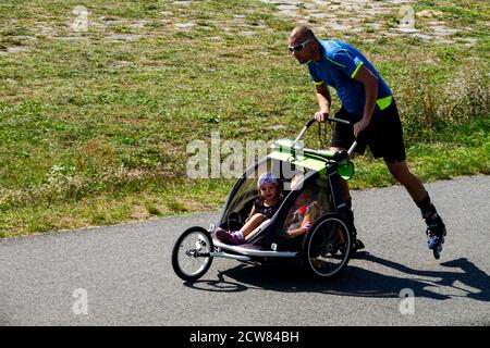 Mann Inline Rollschuhlaufen und schieben Kinderwagen mit zwei Kindern in einem dreirädrigen Buggy-Kinderwagen, laufen Stockfoto