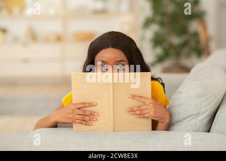 Bleiben Sie zu Hause Hobbys Konzept. Schöne afroamerikanische Frau versteckt hinter Buch auf Sofa drinnen Stockfoto