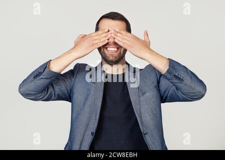 Junger Geschäftsmann Mann mit einem Bart in einer Jacke bedeckt seine Augen mit seinen Händen lächelnd fröhlich und lustig. Blindes Konzept. Porträt eines Mannes auf grauem Hintergrund. Stockfoto
