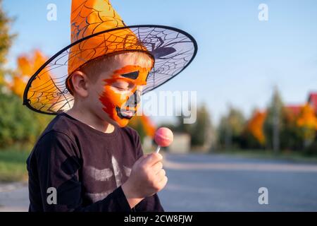 Halloween Kinder. Junge mit Kürbis Gesichtsmaske in Hexenkostüm Hut essen Süßigkeiten aus Eimern sitzen auf der Straße Stockfoto