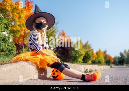 Halloween Kindermaske. Portrait blonde Mädchen im Hexenkostüm mit Kürbis Eimer. Kinder tragen schwarze Gesichtsmasken im Freien Schutz vor COVID-19 Stockfoto