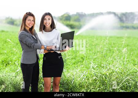 Business-Frauen mit einem Laptop in einem Feld mit Landwirtschaft Bewässerungssystem. Wassersprinkler im Hintergrund. Stockfoto