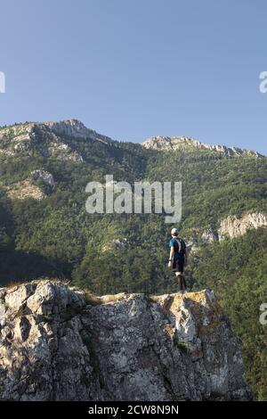 Bergwanderer mit Rucksack, Hut und smarter Uhr, die am Rande einer felsigen Klippe steht und beeindruckende Berggipfel anblickt Stockfoto