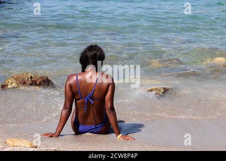 Schwarze Frau in blauen Bikini Sonnenbaden an einem Sandstrand sitzen auf Wellen Hintergrund. Urlaub, Entspannung und Freizeit am Meer Stockfoto