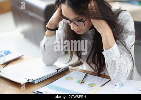 Geschäftsfrau sitzt in einem Büro mit einem Tisch, der ihren Kopf mit den Händen hält und Finanzberichte anschaut. Stockfoto