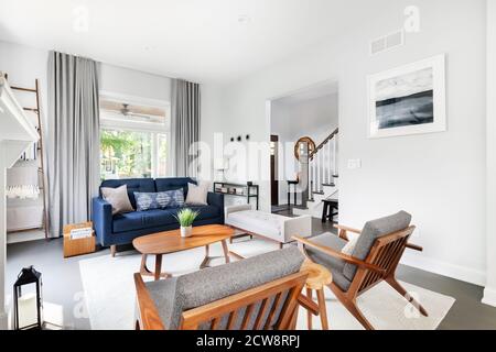 Ein modernes, weißes Wohnzimmer mit Blick auf ein Foyer mit einer Treppe. Eine blaue Couch und zwei Stühle füllen den Wohnraum. Stockfoto