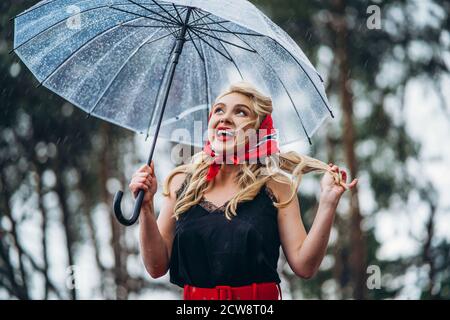 Happy Pin up gestylte Blondine mit roten Lippen zu Fuß in Regnerischer Tag im Freien mit transparentem Regenschirm Stockfoto