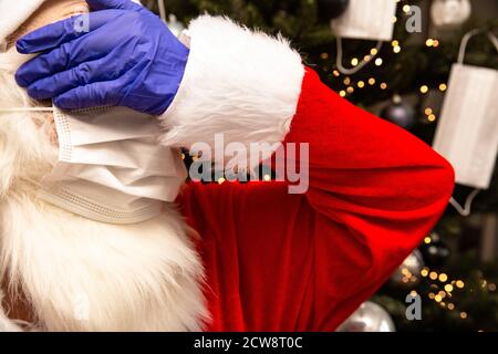 Weihnachtsmann trägt eine Gesichtsmaske und Schutzhandschuhe für Covid-19, Weihnachtsmann steht am Weihnachtsbaum unglücklich über Coronavirus. Stockfoto