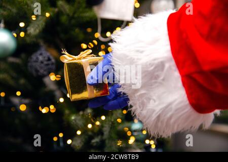 Weihnachtsmann mit Handschuhen und Gesichtsmaske für Coronavirus am Weihnachtsbaum, mit goldenem Geschenk, Covid-19 und Weihnachts-Sicherheitskonzept Stockfoto