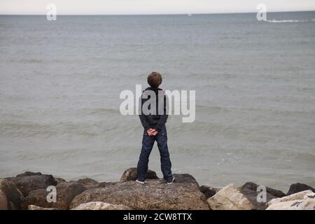 Einjähriger Junge, der auf einer Groyne an der englischen Südküste steht und ein Boot auf See beobachtet Stockfoto