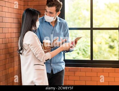 Zwei Geschäftsleute stehen für ein Brainstorming und diskutieren ihre Arbeit mit einem digitalen Tablet. Sie tragen eine schützende Gesichtsmaske im neuen normalen Büro verhindern Stockfoto