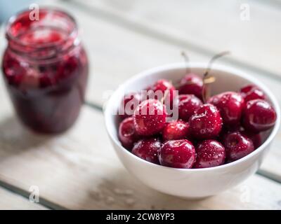 Weiße Keramikschale mit Kirschen und Glas Kirschkonfitüre auf weißem rustikalem Holztisch. Nahaufnahme mit Kopierbereich. Stockfoto