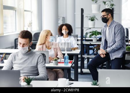 Beratung mit dem Vorgesetzten über Arbeit und soziale Distanz. Millennial Mann und Frau in Schutzmasken reden am Arbeitsplatz im Inneren des Büros Stockfoto