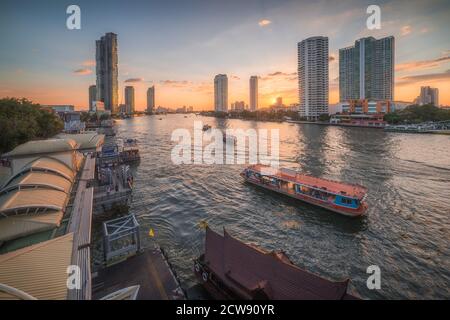 Chao Phraya River mit Wolkenkratzern und Sathon Pier mit Booten bei Sonnenuntergang von der Taksin Brücke in Bangkok, Thailand gesehen Stockfoto