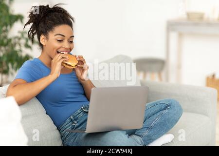Schwarze Frau Genießen Ungesunde Burger Sitzen Auf Laptop Zu Hause Stockfoto