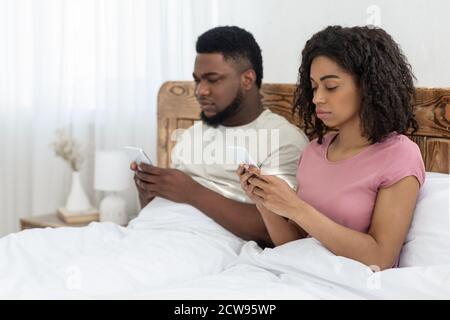 afroamerikanisches Paar sitzt im Bett und benutzt ihre Smartphones Stockfoto