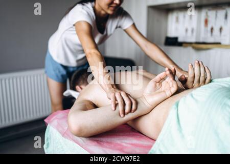 Eine Frau Physiotherapeutin, die eine Rückenmassage für einen Mann in der Arztpraxis macht. Nahaufnahme der Hände, die die Hände des Patienten hinter seinen Rücken legen Stockfoto