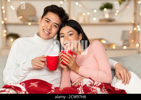 Liebevolle Asiatische Paar Umarmt Holding Kaffee Becher Sitzen Bei Zu Hause Stockfoto