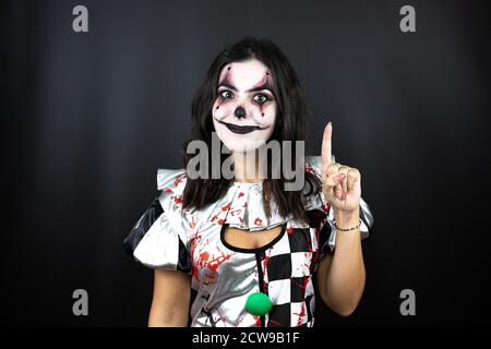 Frau in einem halloween Clown Kostüm über isoliertem schwarzem Hintergrund Zeigen und zeigen mit den Fingern Nummer eins beim Lächeln Selbstbewusst und glücklich Stockfoto