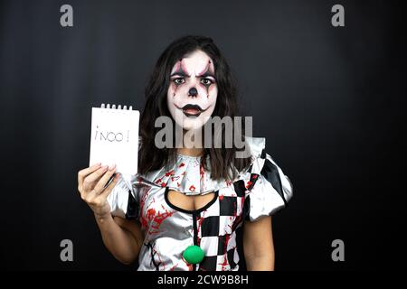 Frau in einem halloween Clown Kostüm über isoliertem schwarzem Hintergrund Erschrocken und überrascht halten Notebook ohne Wort Nachricht Stockfoto
