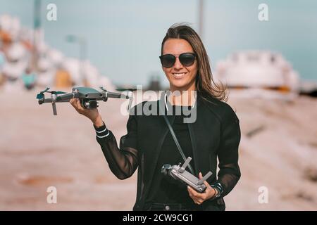 Junge moderne Frau in stilvoller dunkler Kleidung und Sonnenbrille mit Fernbedienung und Drohne vor dem Flug. Stockfoto