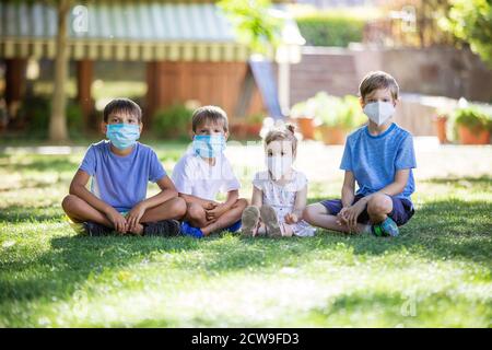 Kleine Kinder in Schutzmasken auf Gesichtern im Freien. Quarantäne. Kinder tragen Sicherheitsmasken, während sie im Park auf Gras sitzen. Coronavirus-Prävention. Stockfoto