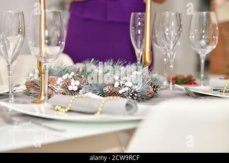 Wählen Sie den Fokus eines Kieferkegelkranzes, umgeben von Gläsern, Tellern und Kerzen auf einem Tisch. Weihnachtsdekoration und Feier Konzept. Stockfoto