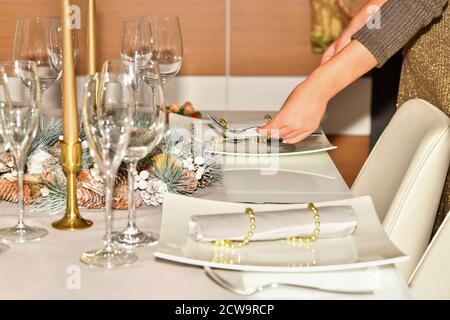 Frau bereitet einen Tisch mit Weihnachtsschmuck, Gläsern und Tellern auf einem unfokussierten Hintergrund vor. Weihnachtsfeier und Dekoration Konzept. Stockfoto