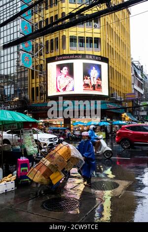 Ein Lieferer mit blauem Plastikregenmantel drückt einen Handwagen, um im Regen in Bangkoks Chinatown Kisten zu liefern. Stockfoto