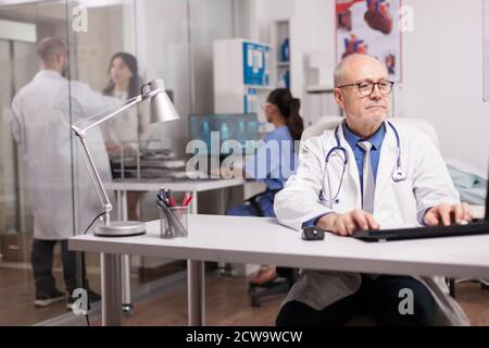 Fokussierter Arzt mit grauen Haaren Typisierung Wissenschaft Bericht über Computer im Krankenhaus Schrank und junge Sanitäter mit weißem Mantel helfen Patienten auf Klinik Korridor. Stockfoto