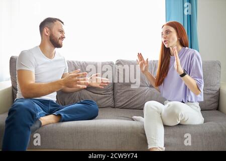 Junge kaukasische Paar wütend auf einander in ihrem Wohnzimmer, schreien einander an, während sie auf dem Sofa sitzen Stockfoto