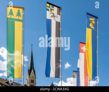 Bunte Fahnen der Schweiz, des Kantons Graubünden und des Val Müstair in Santa Maria, mit dem Kirchturm im Hintergrund Stockfoto