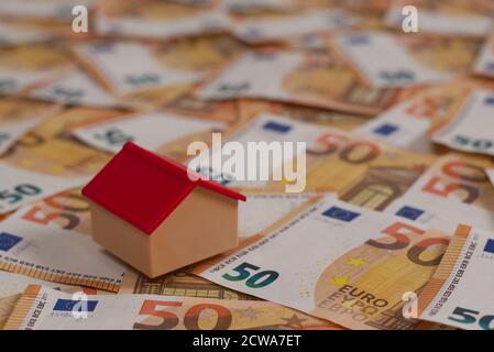 Viele 50-Euro-Banknoten verteilen sich über den Boden, und ein winziges Haus darauf. Darstellung der Kosten für den Kauf eines Hauses. Stockfoto