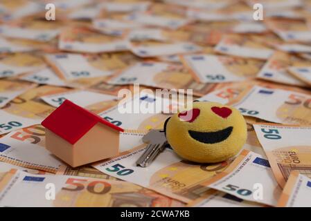 Viele 50-Euro-Banknoten verteilen sich über den Boden, mit einem winzigen Haus und einem Schlüssel an der Haustür. Der Schlüsselhalter ist ein Smiley. Stockfoto