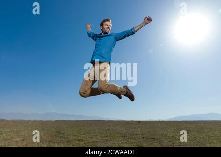 Gut aussehender Casual man trägt blaues Hemd, das mit den Händen in der Luft springt und eine unbeschwerte Haltung draußen am blauen Himmel Stockfoto