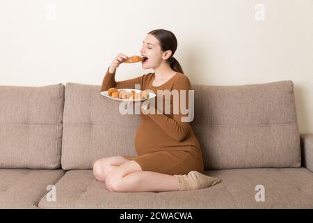 Junge schwangere Frau genießt es, Croissant auf dem Sofa zu essen. Ungesunde Gebäck während der Schwangerschaft Konzept. Stockfoto