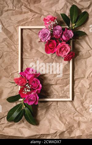 Kreatives Layout mit schönen pinkfarbenen Rosen Blumen und grünen Blättern in Holzrahmen auf zerknittertem Papier. Flaches Lay. Grußkartenkonzept Stockfoto