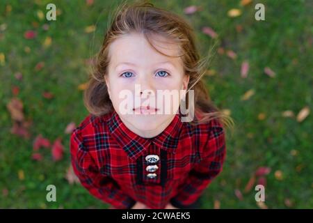 Ein schönes Mädchen mit blauen Augen schaut nach oben. Kind in einem roten Herbstkleid. Blick von oben. Kamera von oben. Schaut in den Himmel. Herbstmotiv Stockfoto