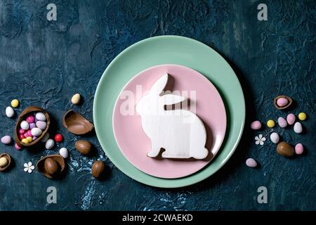 Ostertisch mit Schokoladeneiern, Bonbons und leeren Tellern mit Osterdekoration aus Holz, weißem Hasen. Klassischer blauer Texturhintergrund. Stockfoto