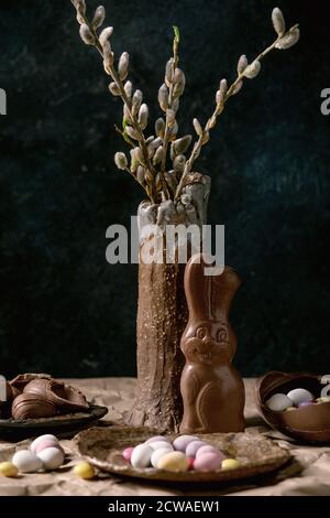 Osterstimmung Stillleben mit Blütenweidenzweigen in Keramikvase, traditionellem Schokoladenkaninchen, Eiern und Süßigkeiten auf dem Tisch mit zerknittertem Bastelpapier. Stockfoto