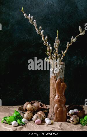 Osterstimmung Stillleben mit Blütenweidenzweigen in Keramikvase, traditionellem Schokoladenkaninchen, Eiern und Süßigkeiten auf dem Tisch mit zerknittertem Bastelpapier. Stockfoto